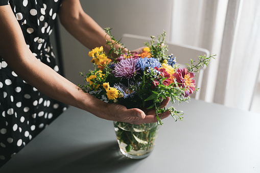 young Caucasian woman with black dress and white polka dots arranging a bouquet of freshly picked up beautiful flowers in a glass vase on a grey table in natural day light