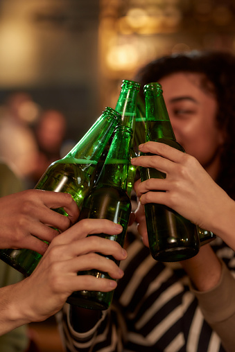 Good vibe for good night. Close up of hands of people in the bar clinking bottles while drinking beer together. Friendship and entertainment concept. Focus on hands and bottles. Vertical shot