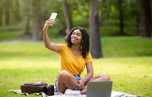Cool black girl with laptop computer taking selfie during her outdoor studies at park, panorama