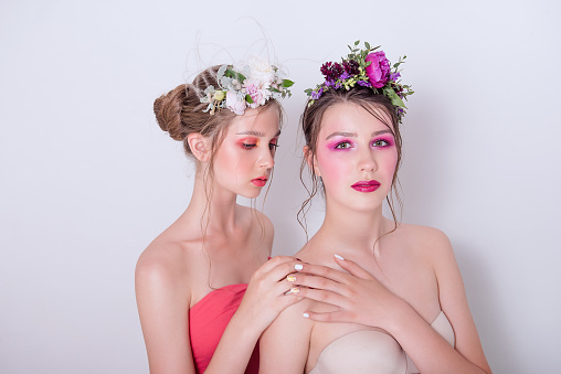 Two fashion beauty models sister girls with professional bright make-up, a hoop of fresh spring flowers on their heads, with red lilac lipstick are posing against a gray uniform background. Clean skin