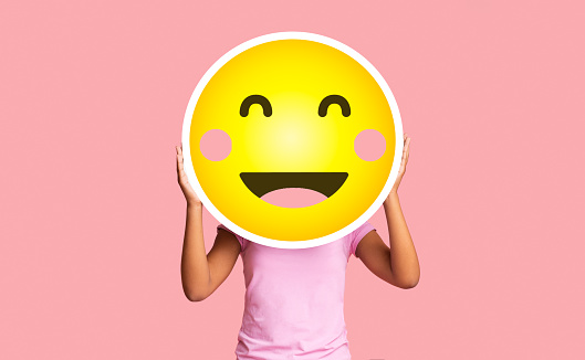 Black girl hiding face behind happy shy emoji emoticon, standing over pink studio background with copy space