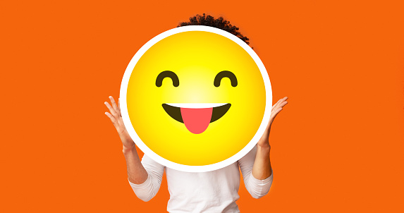 Black bushy man holding happy cheeky emoji emoticon smiley in front of his head, standing over orange studio background, creative collage, empty space