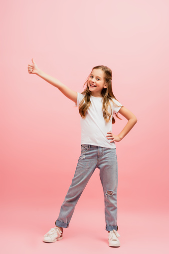 child in white t-shirt with hand on hip showing thumb up while looking at camera on pink