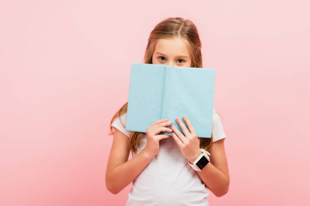 istock child in smartwatch obscuring face with book while looking at camera isolated on pink 1269076006
