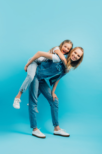 full length view of young woman in denim vest and jeans piggybacking daughter while looking at camera on blue