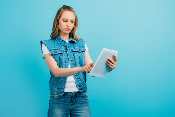 concentrated woman in denim vest using digital tablet isolated on blue