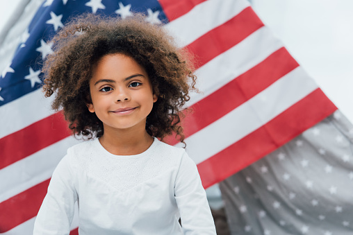 curly african american kid looking at camera near flag of america