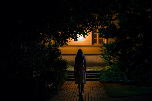 One young alone woman in dress walking on sidewalk through dark park to home in summer black night. Scary moment and gloomy atmosphere. Back view.