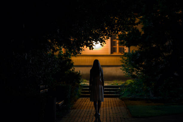 одна молодая одинокая женщина в платье гуляет по тротуару через темный парк к дому в летнюю черную ночь. страшный момент и мрачная атмосфер� - street alley dark city стоковые фото и изображения