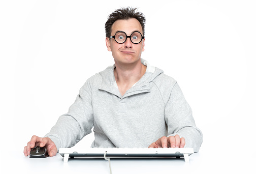 Surprised man with glasses sits at the keyboard in front of the computer, on white background. Funny programmer