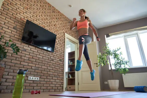 Fit sporty active young adult woman wearing sportswear doing fitness sport exercise at home. Athletic strong girl jumping up in air indoor in modern apartment living room. At home gym workout concept.