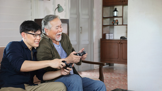 Senior Asian man in retirement age having fun playing video game console with his son in his cozy home with copy space