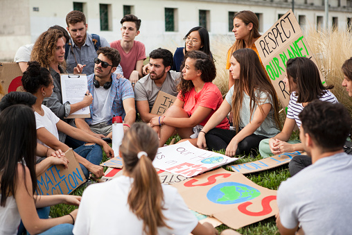 Group of young male and female protestors discussing over clipboard while sitting outdoors