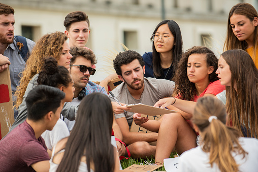 Group of young male and female protestors discussing over clipboard while sitting outdoors