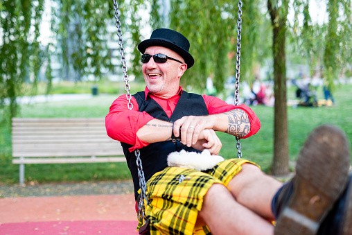 eccentric and crazy man playing on a children's swing, man wearing sunglasses and bowler hat in a playful pose, original clothing with red shirt, waistcoat with pins and Scottish kilt