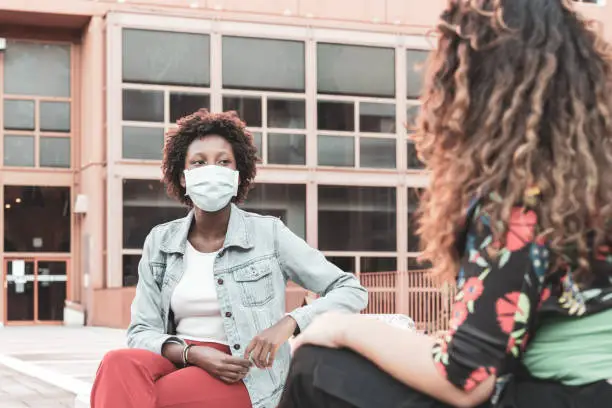 Photo of two young women sitting on a bench, chatting using the protective mask and the social distancing, multiracial couple of friends, filter effect vintage