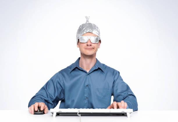 conceptual man with an aluminum foil hat and aluminum glasses behind a keyboard in front of a computer. - tin foil hat imagens e fotografias de stock