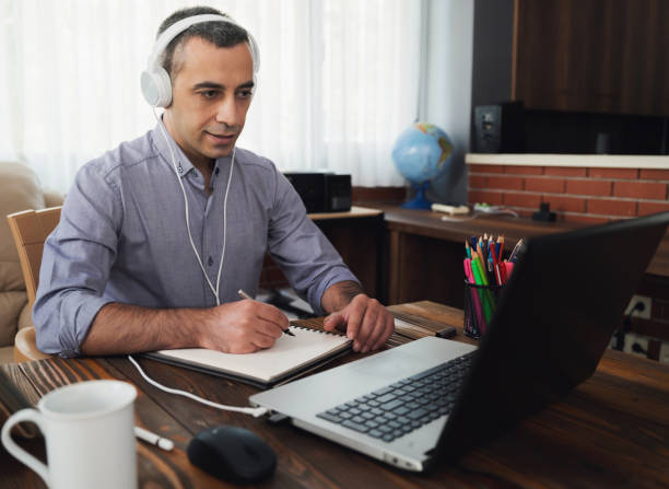 istock Man Is Working Online In Home Office Using His Laptop Computer, Joining Webinar And Taking Notes On Digital Tablet 1269145470