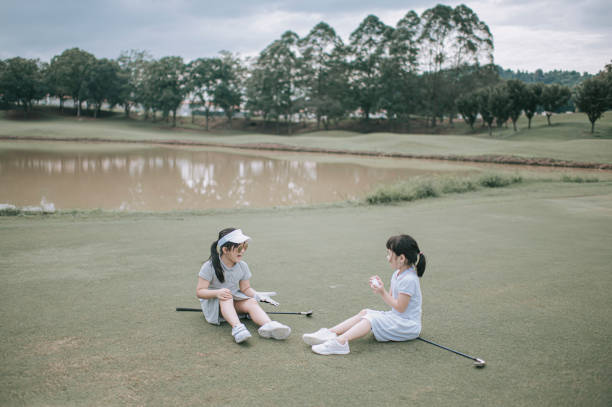 2 asian chinese young girls sitting on the green waiting for their parent in golf course during weekend - golf course golf people sitting imagens e fotografias de stock