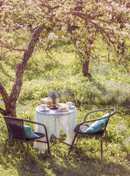 Springtime in garden. Table set with tea cups, boho reuse jar vase with forget me not flowers( Myosotis) and small stack of books. Summer relax readings concept.