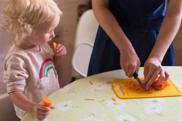 petite fille avec les cheveux bouclés blonds soutenant sa cuisine de mère dans la cuisine - baby carrot photos et images de collection