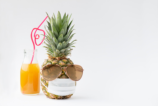 A bottle of fruit juice and a pineapple wearing sunglasses and protective face mask in front of white background. Representing holidays in lockdown situations.