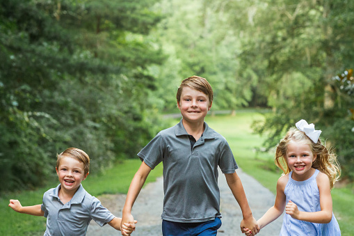 Three sibling children running outside on a warm summer day down a long country tree lined road