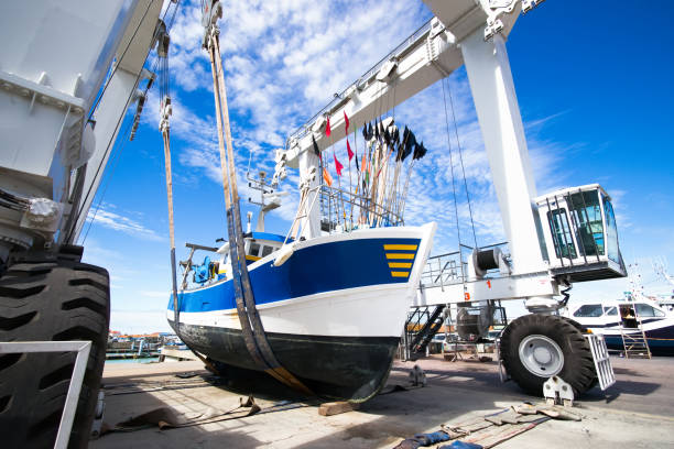 Wide angle photo of fishing boat lifted by a boat lift. Blue and white boat, white boat lift and blue sky. A fishing boat is suspended for maintenance. Photo from front right. dry dock stock pictures, royalty-free photos & images