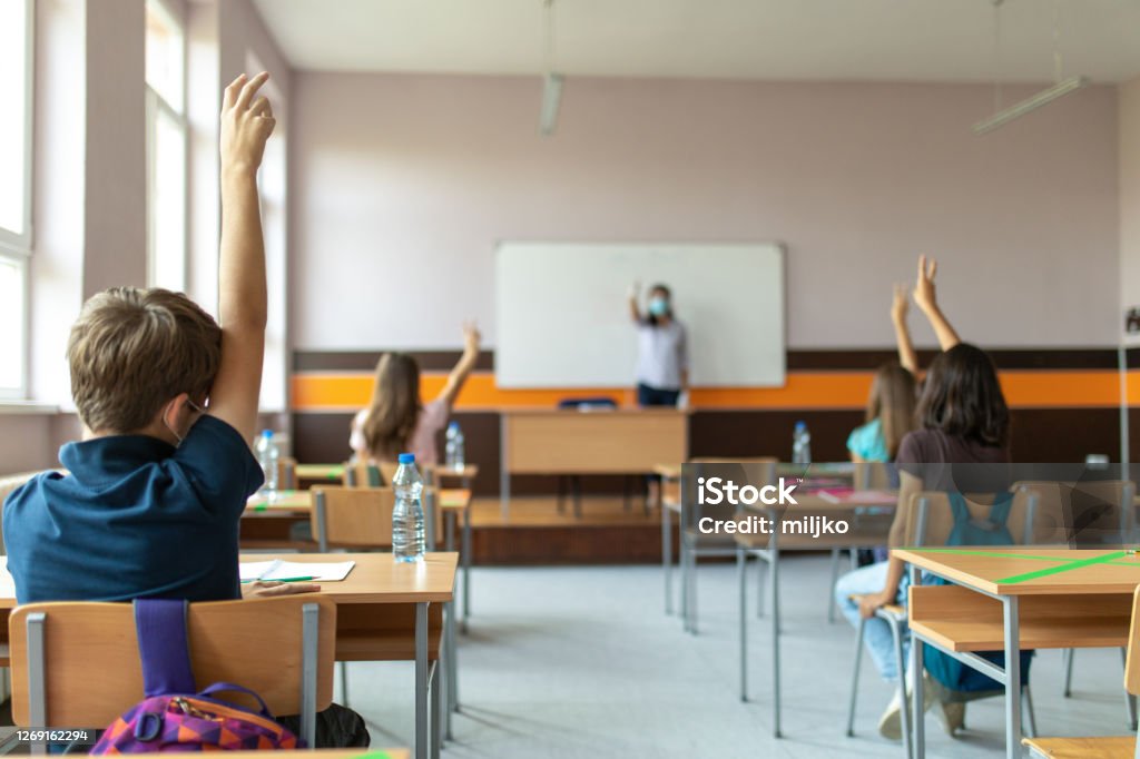 Back to school Students with protective masks sitting in school desks in their classroom. School desks are marked with a cross to mark a place where sitting is not allowed to maintain social distance during corona virus . Education Stock Photo