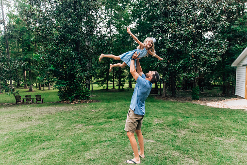 a daddy playing outside with his daughter by lifting her up in the air like she is flying