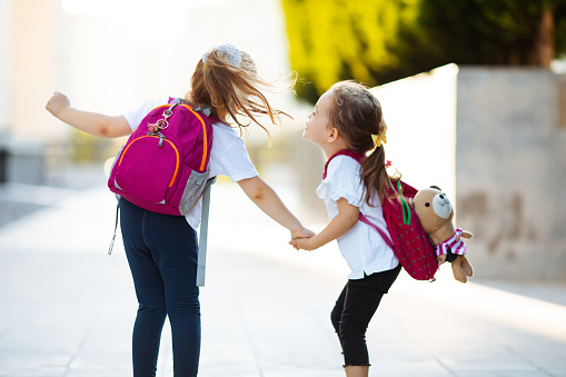 Portrait of smiling little pretty girls holding hands , wearing backpacks and walking to school.
