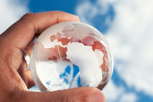 Mans hand holding a world globe. There is blue sky in the background. The globe is made of glass. Close up