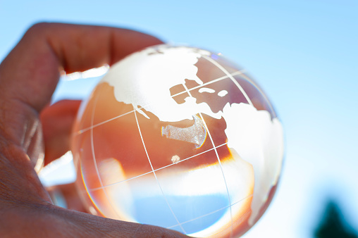 Mans hand holding a world globe. There is blue sky in the background. The globe is made of glass. Close up