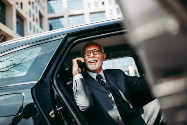 Good looking senior business man sitting on backseat in luxury car. He opens car doors and going or stepping out. Big busness bulding in background. Transportation in corporate business concept.