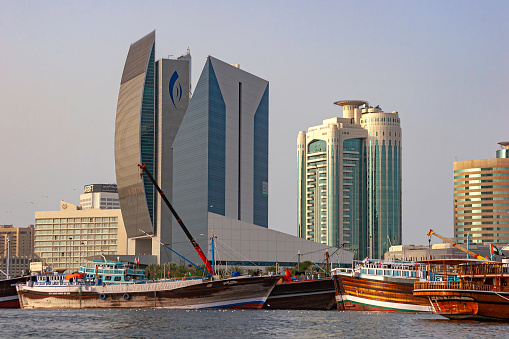 Dubai, United Arab Emirates - December 28, 2006: Traditional Arab dhows, moored on the Deira side of Dubai Creek. They are waiting to be loaded. It is from here that local arabs plied their shipping trade to India and the East Coast of Africa, for several hundreds of years.  \nIn the background are some modern office buildings on the waterfront. Photo shot from a boat on the Creek, in the late afternoon sunlight; horizontal format. Copy space.