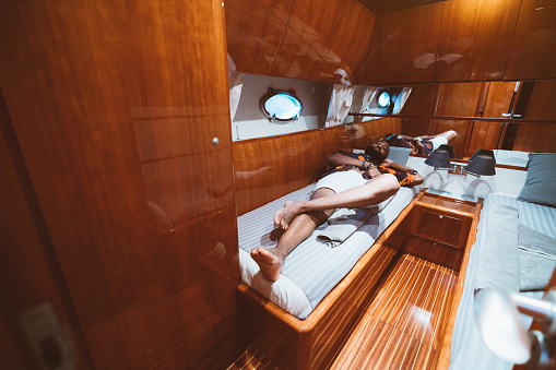 A rich carefree bearded black guy in shorts is laying on a bed in a cabin of a luxury yacht with a porthole, beds, reflective wooden walls and a mirror; an African tourist guy relaxing in a boat cabin
