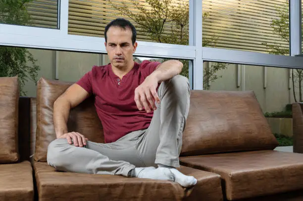 Photo of Sad and downcast Brazilian mature man (44 years old), sitting on the brown sofa, with a large window pane behind.