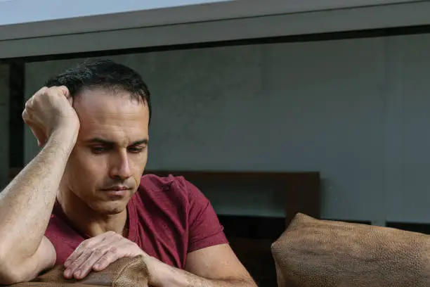Photo of Mature Brazilian man (44 years old) sitting on the brown sofa, behind the glass, sad and crestfallen.