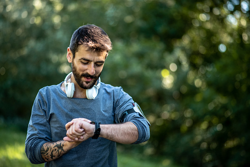 A young man stands in nature and looks at his smart watch on a sunny day