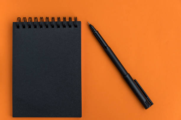 istock Black blank notepad and pen on an orange background. Minimalistic halloween celebration design. Mock up. Copy space. 1269243125