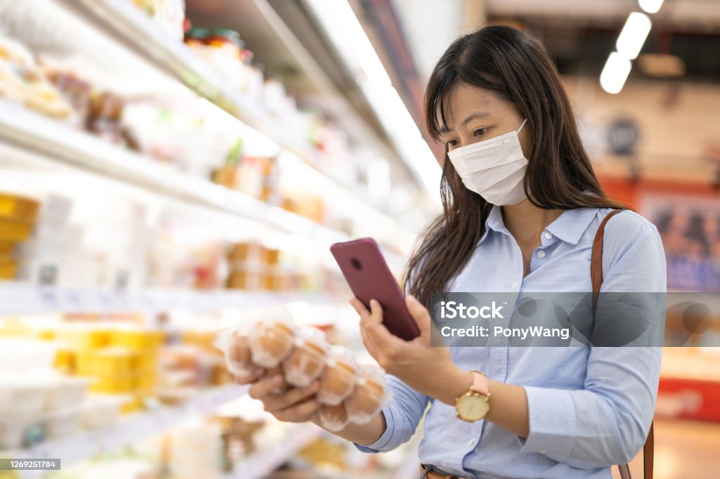 businesswoman picks up eggs asian young businesswoman wear face mask who is shopping for fresh eggs and scanning barcode with smart phone at supermarket Medical Scan Stock Photo