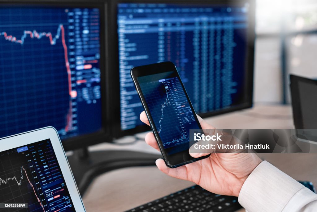 Stocks and Funds. Trader sitting at office in front of monitors with data using app on smartphone monitoring price changes close-up Man trader sitting at desk at office in front of monitors with stocks data holding smartphone browsing application monitoring price changes on candle bar online using digitla devices close-up Bitcoin Stock Photo