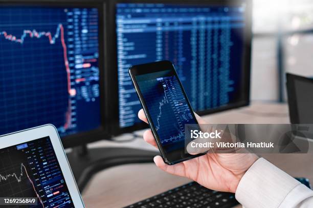Stocks And Funds Trader Sitting At Office In Front Of Monitors With Data Using App On Smartphone Monitoring Price Changes Closeup Stock Photo - Download Image Now