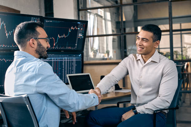 Trading Concept. Manager hiring intern shaking hands cheerful sitting at office in front of monitors with price flow charts