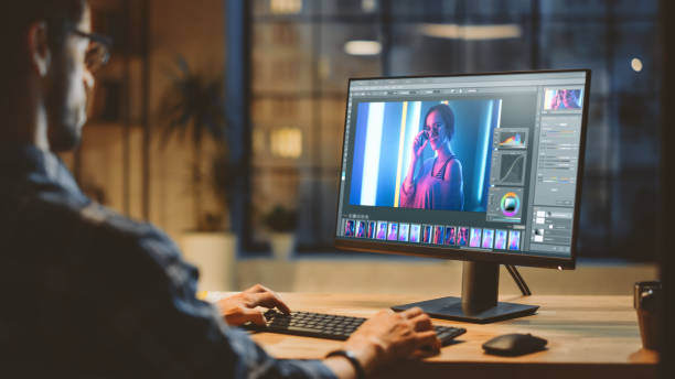 istock Over the Shoulder: Creative Young Digital Editor Works in Photo Editing Software on His Personal Computer with Big Display. In the Background Evening in Modern Loft Studio Office 1269257031