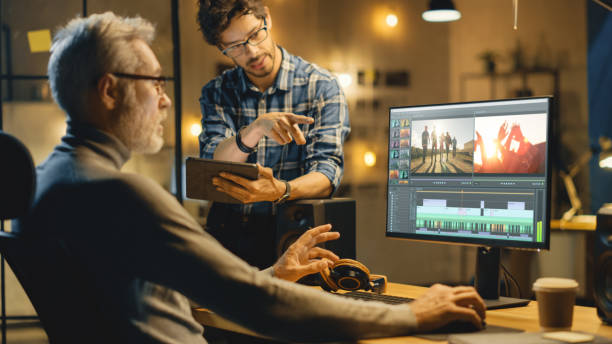 istock Evening in Creative Office: Professional Middle Aged Videographer Works on Desktop Computer, Has Conversation with Younger Specialist who Uses Tablet Computer. Modern Video Developers Office 1269257044