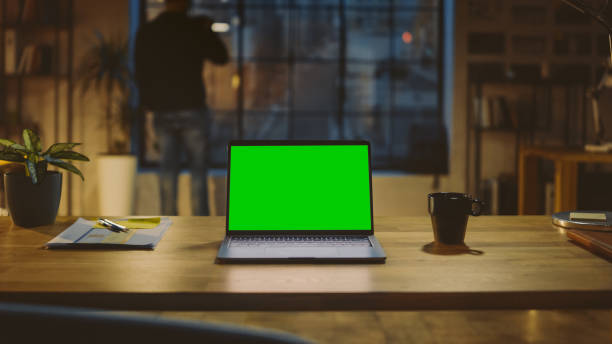 istock Desktop Computer with Mock-up Green Screen Standing on the Wooden Desk in the Modern Creative Office. In the Background Designer Drinks from a Cup Looks at the Night City out of the Window 1269257080