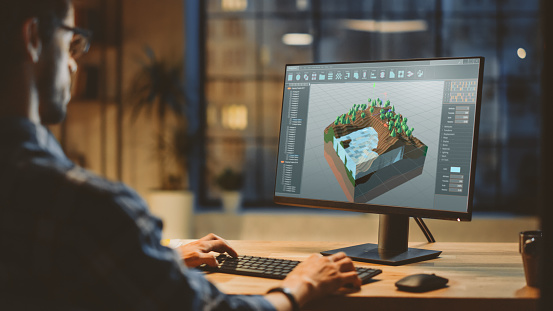 Por la noche, Creative Young Video Game Developer funciona en un ordenador de sobremesa con diseño de nivel de videojuego 3D de pantalla. Elegante Creative Studio Office. Over the Shoulder Shot photo