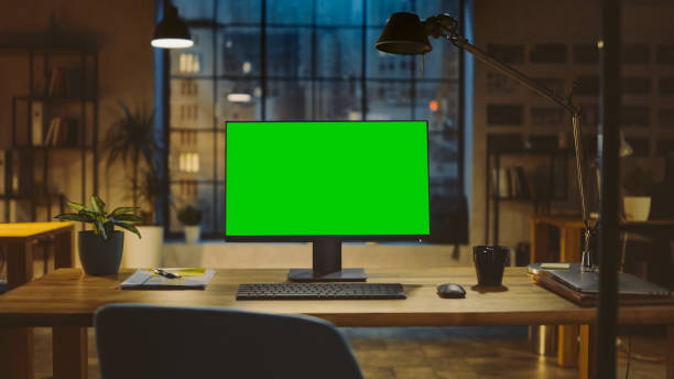 istock Desktop Computer with Mock-up Green Screen Standing on the Wooden Desk in the Modern Creative Office. In the Background Warm Evening Lighting and Open Space Studio with City Window View. 1269257100