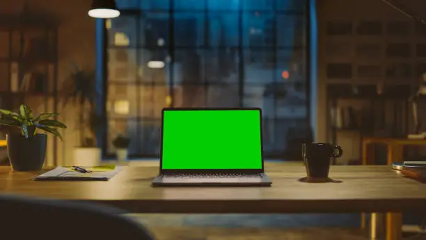 Mock-up Green Screen Laptop Standing on the Desk in the Modern Creative Office. In the Background Warm Evening Lighting and Open Space Studio with City Window View.